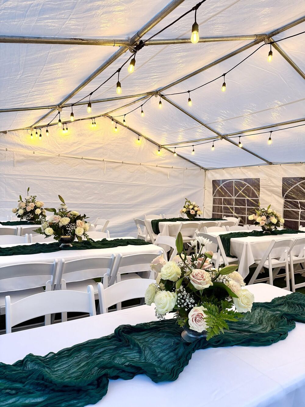 String lights glowing under a white tent with tables, green runners and flowers